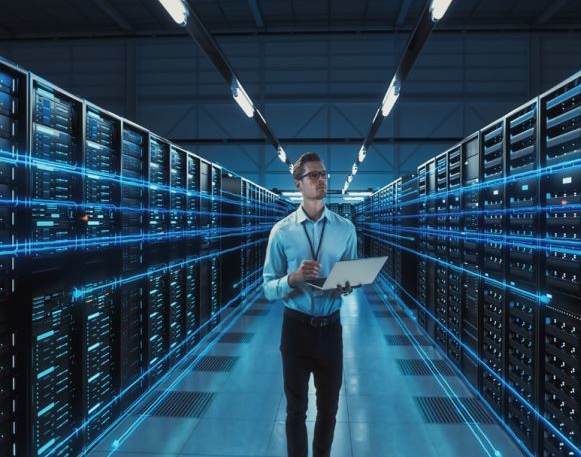 Man in a blue button-up shirt stands in a server room holding an open laptop