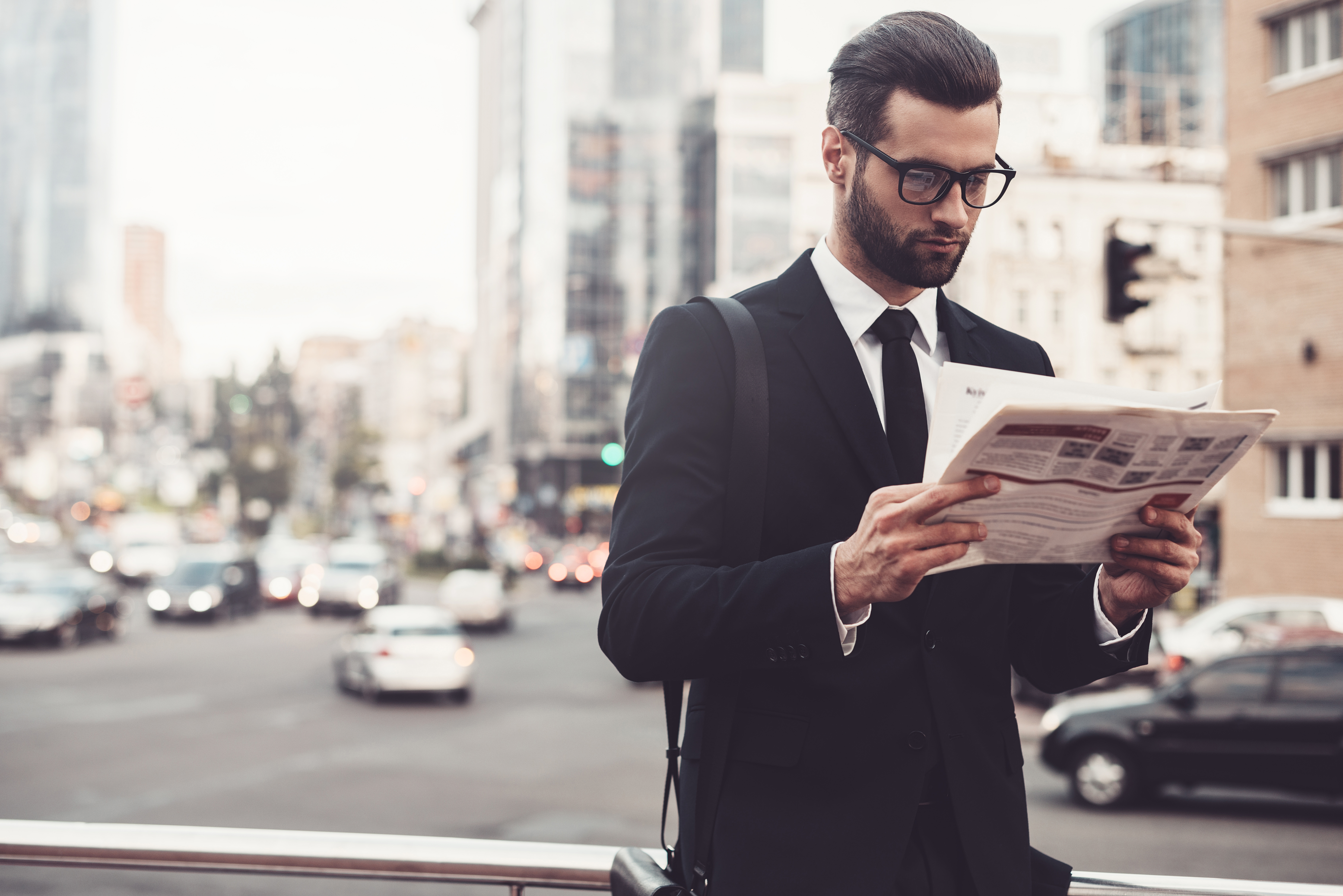 Man in a suit and sunglasses, reading a newspaper outside on the street in a US city.